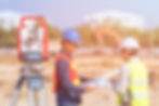 Surveying equipment on tripod in focus; two construction workers with helmets and vests examining plans in a blurred construction site.