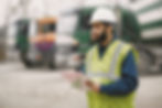 Construction worker in a hard hat and safety vest holds a clipboard at a work site, with blurred trucks behind him.