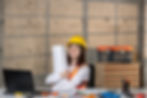 Engineer in a yellow helmet and orange vest smiles, holding blueprints. Desk with blueprint tools, concrete wall, toolbox in the backdrop.
