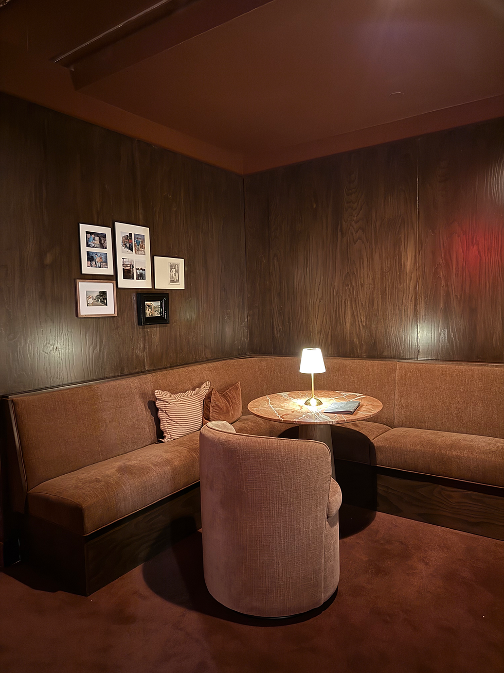 2026 interior design trends. Cozy brown seating area with corner couch, round table, lamp, and framed photos on wood-paneled walls. Warm and inviting ambiance.