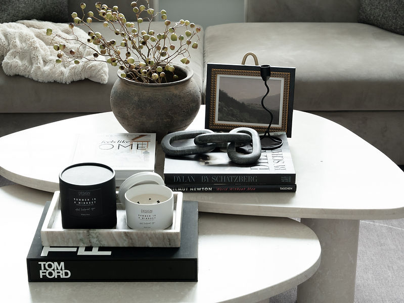 Styled vignette in Project Pheabs featuring dark vase, candles, books, black chunky chain link, curved candlestick, landscape artwork, and grey couch in a cozy, globally inspired living area.
