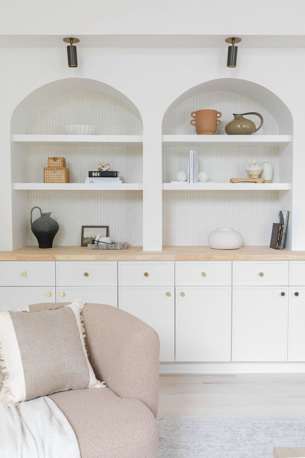 Modern living room with white shelves holding pottery and books, a beige sofa with a cushion, and minimalistic decor. Bright, clean, cozy.