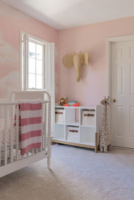 Serene and tranquil pink nursery with cloud wallpaper, white crib, striped blanket, rattan elephant, giraffe plush, and storage details.