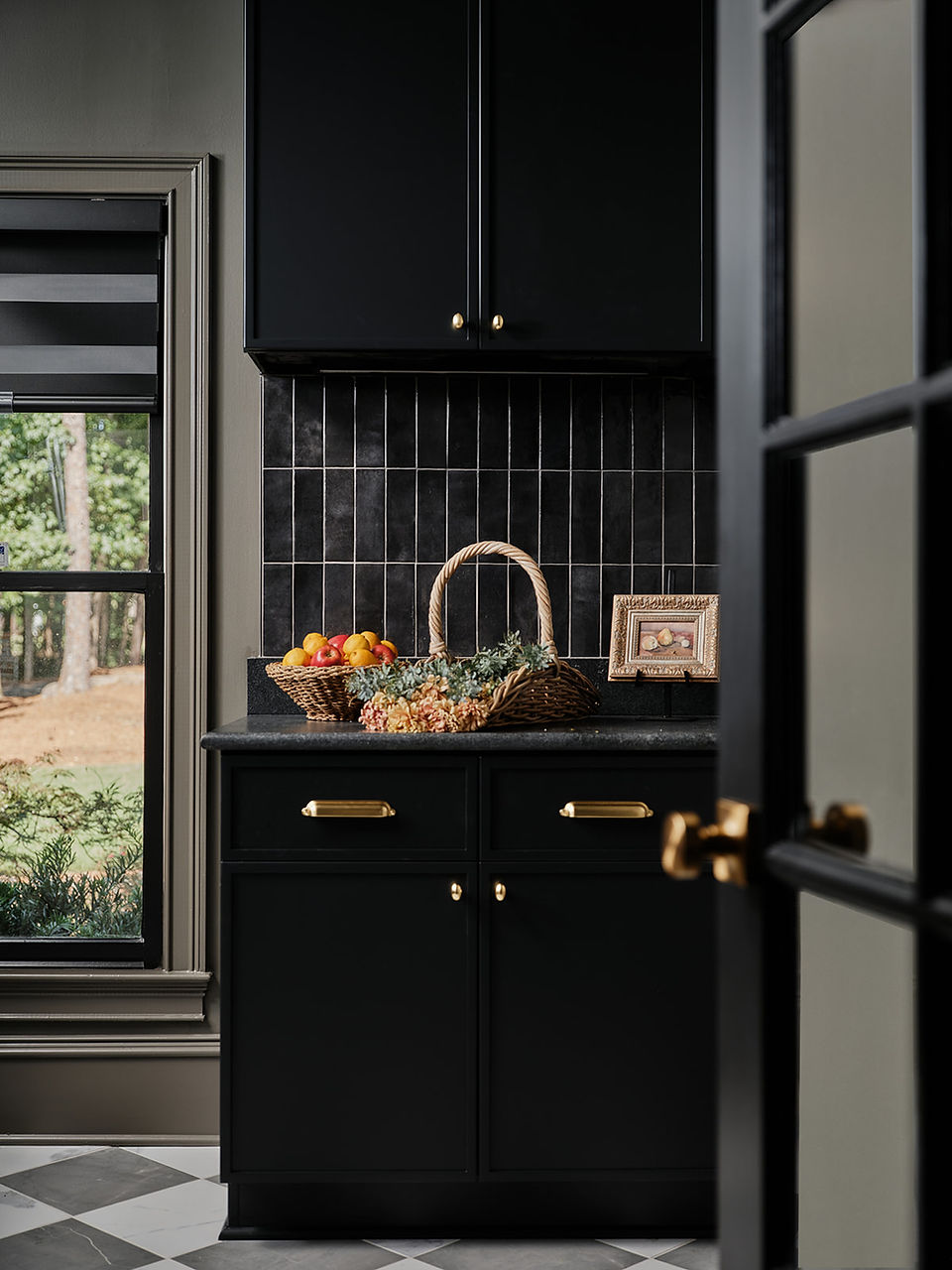Moody black kitchen nook with gold hardware, woven fruit baskets and flowers on the counter beside a window.