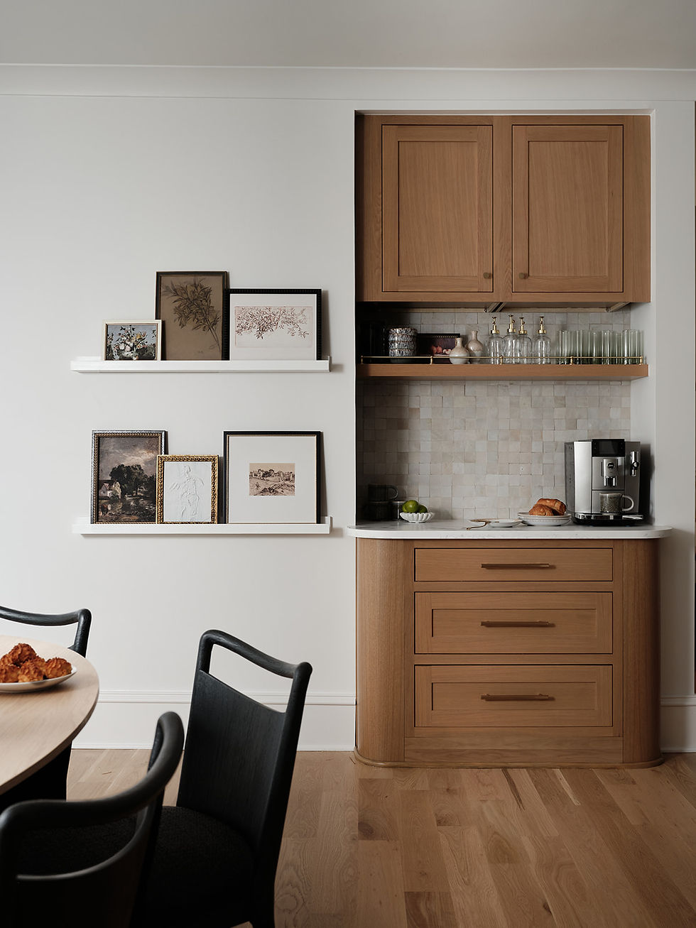 Modern kitchen with wood cabinets, framed art on white shelves, coffee machine, pastries, and black dining chairs in a calm setting