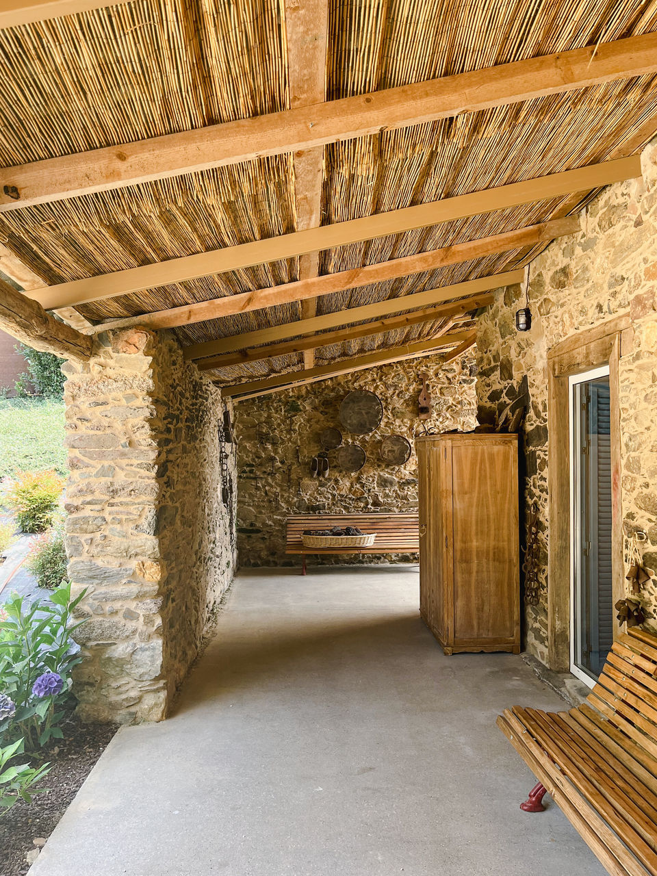Porche de acceso que rodea la casa. Muros de piedra, falso techo de cañizo, banco de madera y hierro forjado