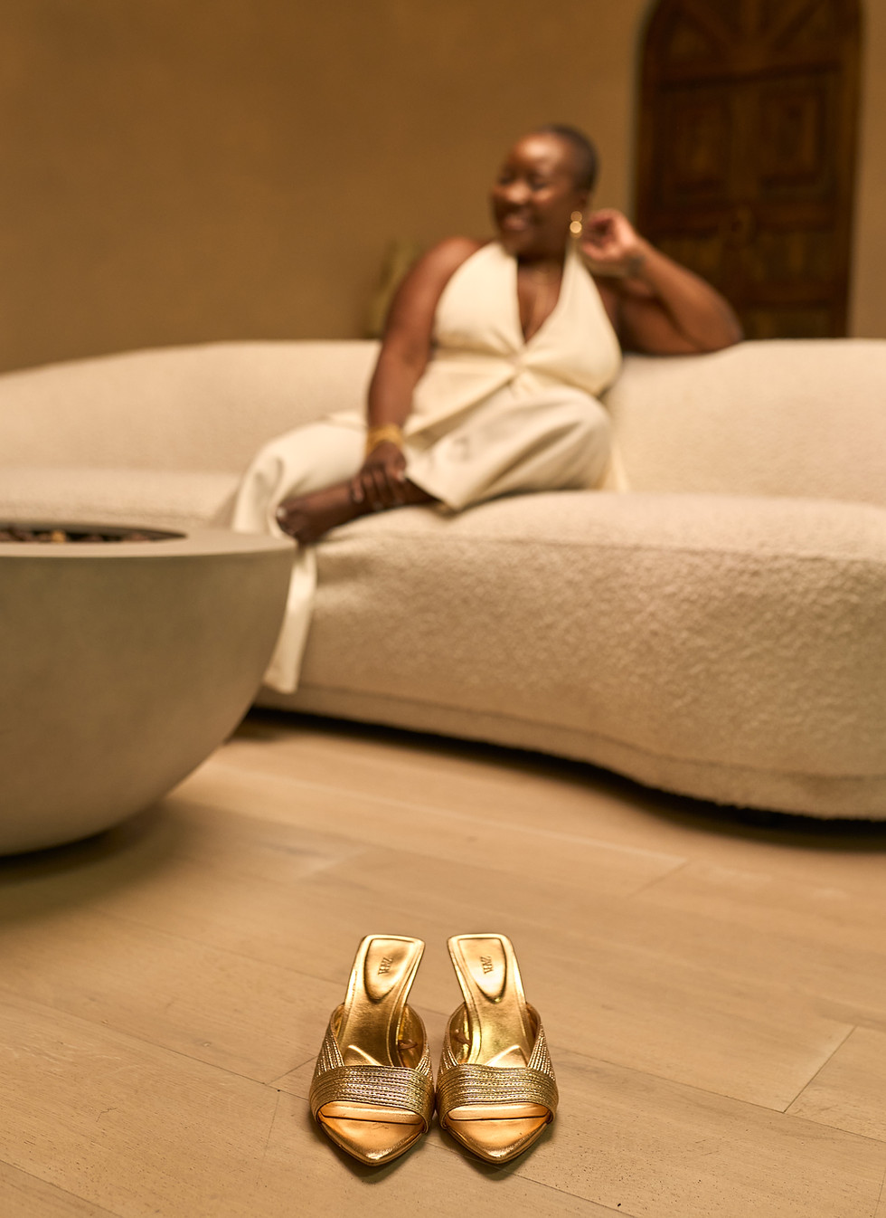 Nubia Younge smiling while sitting on a couch in a white jumpsuit, with gold flats in the foreground.