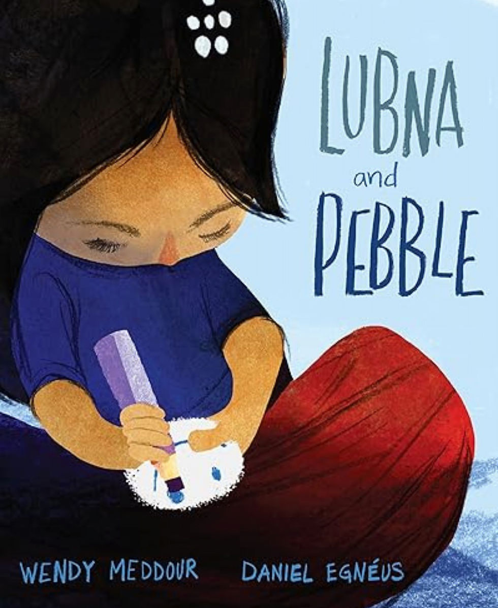 A child in blue colors a pebble on a red background. Text: Lubna and Pebble, Wendy Meddour, Daniel Egnéus. Serene mood in a refugee camp.