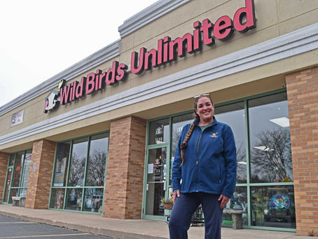 Smiling woman in blue jacket stands outside "Wild Birds Unlimited" store, with brick facade and large windows displaying merchandise.