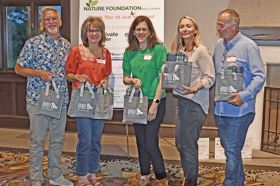 Five people with gift bags stand indoors in front of a "Nature Foundation" sign. They appear cheerful, wearing casual attire.