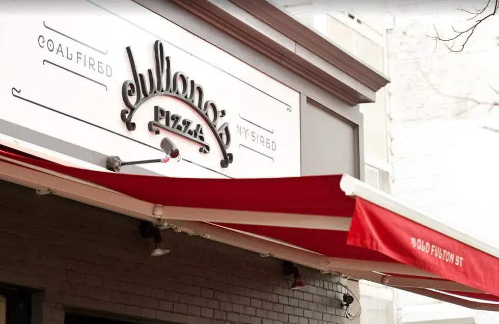 Red and white striped awning outside a pizzeria. Sign reads "Juliana’s Pizza." Brick building exterior. Text: "Coal Fired" and "19 Old Fulton St."