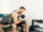 Man sitting on a bench lifting a dumbbell in a home gym. He wears a green tank top and black shorts. Weights and a sofa in the background.