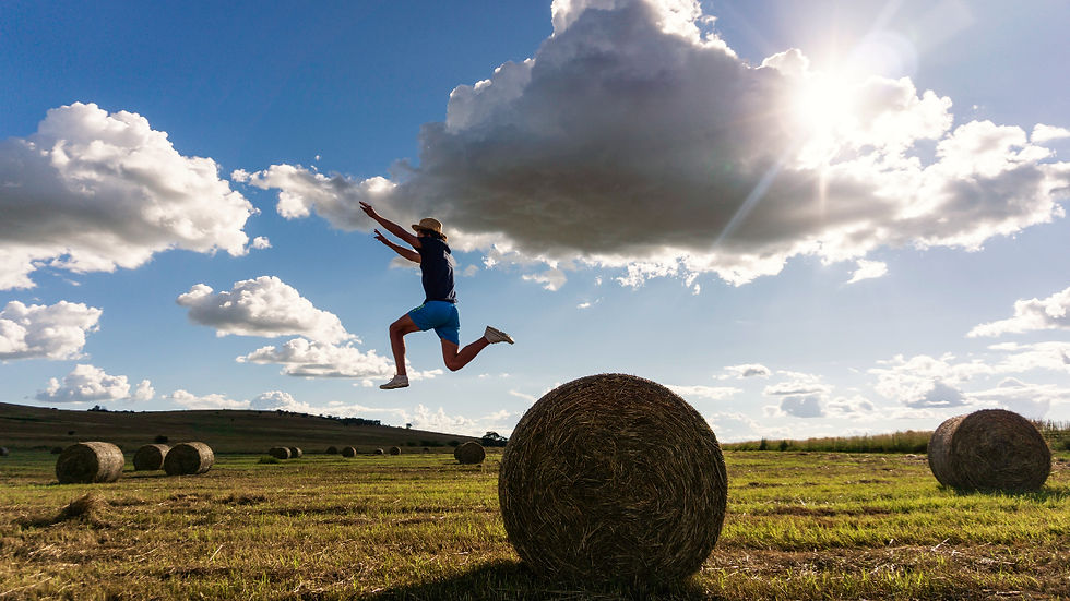 Happy person jumping over hay bales in a sunny field, wearing blue shorts and a hat, showing stress free life.