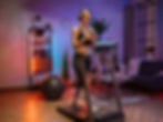 Women jog on a treadmill in a cozy room with neon lights, wearing headphones and holding phones. A yoga ball and plants are in the background. Click on the image to go to the homepage.