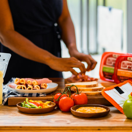 Person preparing sandwiches on a wooden table with tomatoes, lemonade, sliced cheese, and bread. Bright, casual kitchen setting.