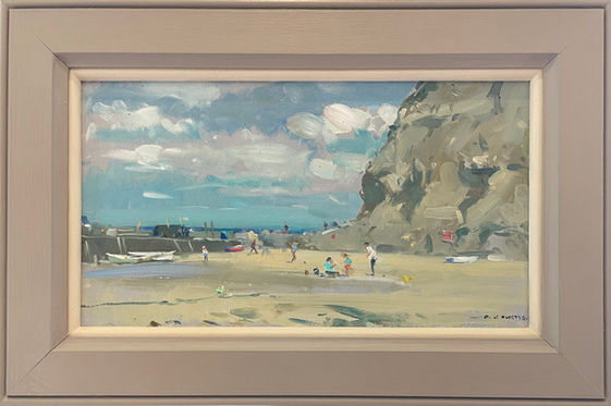 A view of people enjoying the sandy beach of Staithes harbour with a glimpse of the rock armour and cliff face, pink clouds scudding across the blue sky in the background.