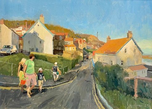 A group of holiday makers walk along the road towards the charming coastal cottages of Runswick Bay gleaming in the late afternoon sun.