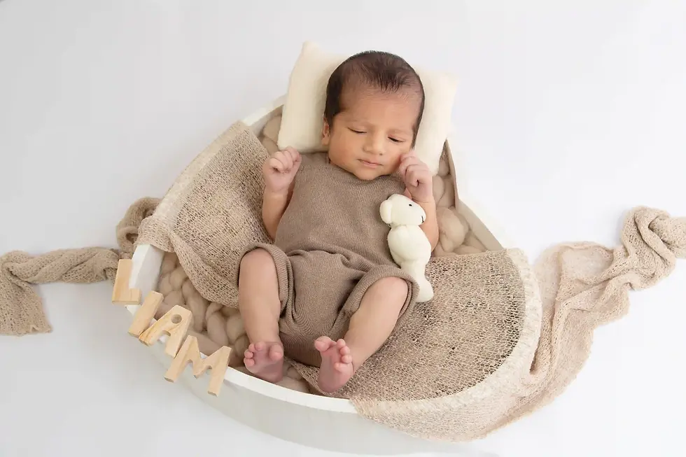 Schlafendes Baby im beigen Strampler in weißer Korbwiege mit Teddybär; Holzbuchstaben Liam auf hellem Hintergrund.