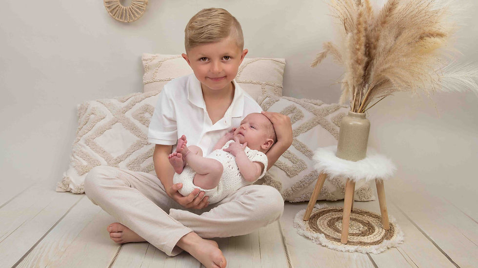 Junge im weißen Outfit hält ein Baby auf dem Schoß, sitzt vor beigem Bett mit Kissen und Trockenblumen, ruhig und liebevoll.