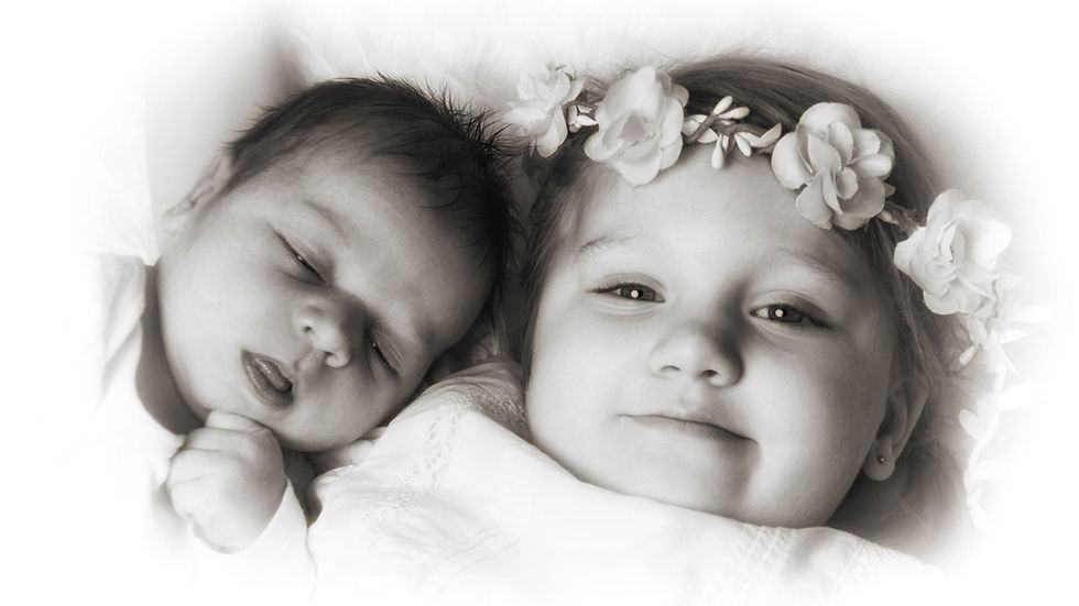 Schlafendes Baby neben kleinem Kind mit Blumenkranz, beide auf weißer Decke, zartes Schwarzweiß-Porträt.