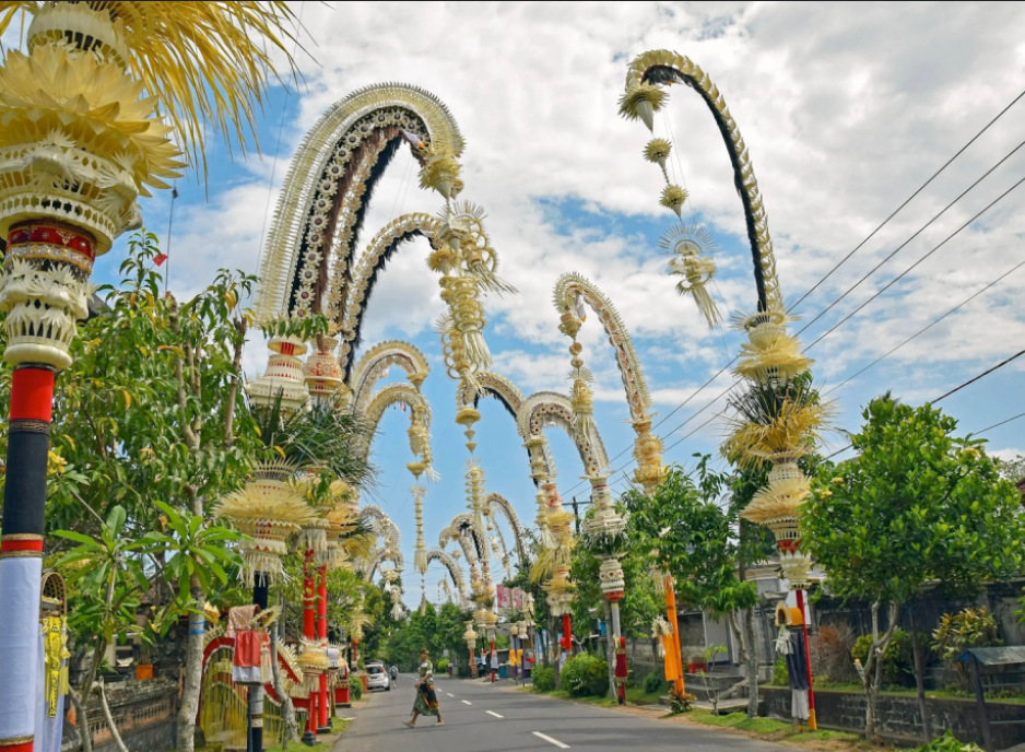 Un boulevard bordé d'arbres à Bali, décoré de plusieurs grands et élégants penjor (arcs en bambou) aux ornements blancs et dorés, sous un ciel bleu nuageux.