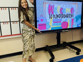A teacher smiles and motions to a smart board that has a message welcoming students to kindergarten.