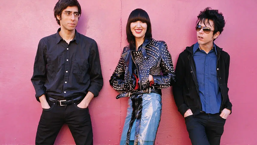 Retrato promocional de la banda de indie rock Yeah Yeah Yeahs en Nueva York en 2003 para el lanzamiento de su álbum Fever to Tell, con Brian Chase, Karen O y Nick Zinner posando frente a una pared rosa.
