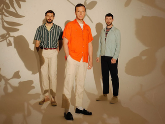 Integrantes de la banda Friendly Fires posando en estudio. Ed Macfarlane en el centro con camisa naranja brillante, flanqueado por Edd Gibson y Jack Savidge, frente a una pared iluminada con sombras de plantas.