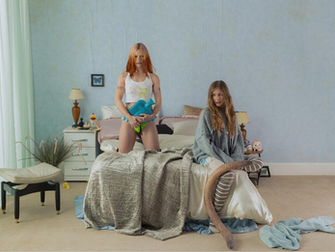 Fotografía promocional de la banda Wet Leg en una habitación surrealista con paredes azul claro. Una integrante posa de pie sobre la cama con ropa blanca y una bolsa de agua caliente azul, mientras la otra se sienta al borde del colchón junto a un objeto con forma de gusano gigante