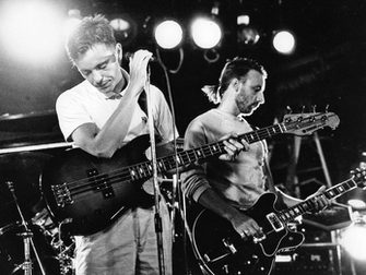 Fotografía en blanco y negro de los músicos Bernard Sumner (izquierda, cantando al micrófono) y Peter Hook (derecha, tocando el bajo), miembros de la banda británica New Order, actuando en vivo en la discoteca Paradise Garage en Nueva York el 7 de julio de 1983