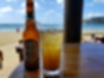 Bottle and glass on a wooden table, with a straw in the drink. Sandy beach and ocean in the background, under a clear blue sky. Relaxed mood.