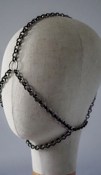 Black and silver chain headpiece displayed on a fabric mannequin head, with linked chains crossing over the crown, forehead, sides of the face, and under the chin.