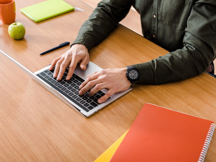Person typing on a laptop at a wooden desk with a green notebook, red folder, apple, pen, and orange mug. Wearing a dark watch.