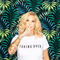 A picture of Sam Divine with her finger to her lips against a tropical leafed background.
