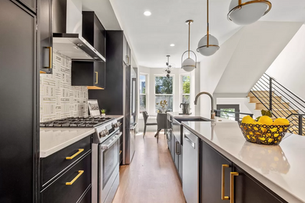 Modern kitchen with black cabinets, gold accents, and patterned backsplash. Sunlit dining area in background. Bowl of lemons on counter.