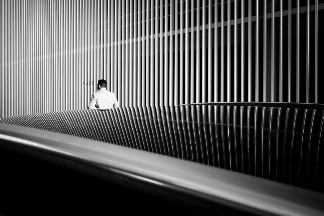 Minimal black-and-white photograph of a person walking through a dense pattern of vertical lines, creating rhythm, repetition, and depth in an urban architectural setting.