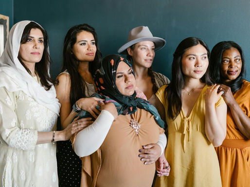 Diverse group of six women standing together against a blue wall, some holding hands, wearing colorful outfits, with a calm, supportive mood.