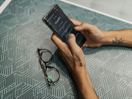 Hands holding a smartphone displaying "ChatGPT" over a teal patterned table. Glasses lie beside, tattoos visible on wrists.