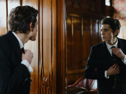 A man in a suit adjusts his tie while looking in a mirror, set in a wood-paneled room with vintage decor.