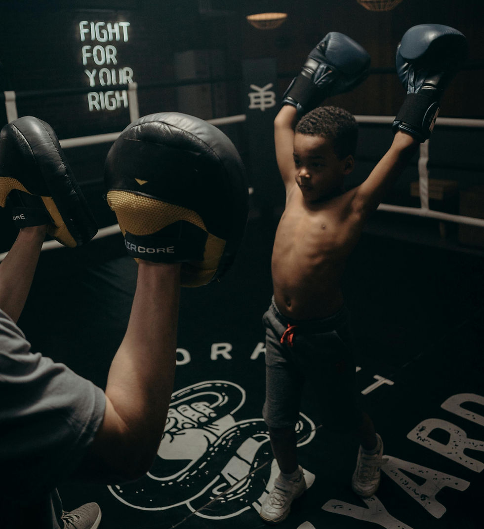 Young boy in boxing gloves raises arms triumphantly in a dimly lit gym. Trainer holds pads. Neon sign: "FIGHT FOR YOUR RIGHT."