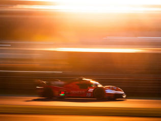 Carro de corrida vermelho percorre uma pista em alta velocidade durante o pôr do sol. O efeito de arrasto das luzes e o fundo desfocado destacam a velocidade do veículo, enquanto a iluminação dourada cria uma atmosfera intensa e dramática típica de competições de endurance.