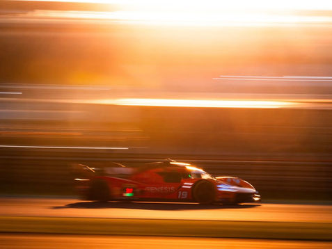 Carro de corrida vermelho percorre uma pista em alta velocidade durante o pôr do sol. O efeito de arrasto das luzes e o fundo desfocado destacam a velocidade do veículo, enquanto a iluminação dourada cria uma atmosfera intensa e dramática típica de competições de endurance.