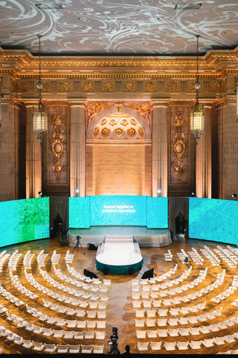 A dynamic event setup designed for thought-leadership presentations and keynote addresses. The stage features a sleek  circle centered under professional lighting, flanked by digital screens and minimalist décor. Audience seating is theater-style, emphasizing sightlines and engagement. The overall aesthetic blends the gravitas of the historic venue with the energy of modern ideas, perfect for conferences, speaker series, and innovation summits
