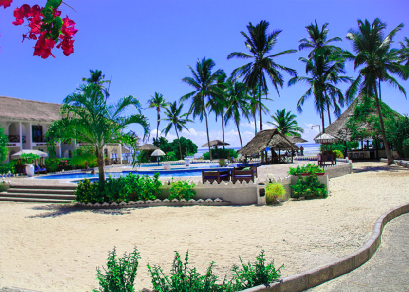 Tropical beachfront resort courtyard with palm trees, flowering plants, and a sandy pathway leading