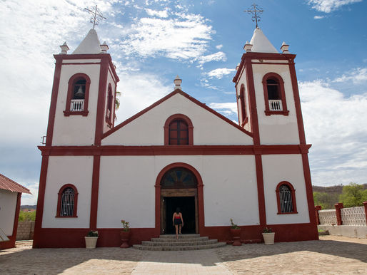 El Triunfo Church in Baja California Sur stands prominently under a sunny sky, with its distinctive red and white facade and twin bell towers. A visitor approaches the entrance, adding a sense of scale to this historical landmark.
