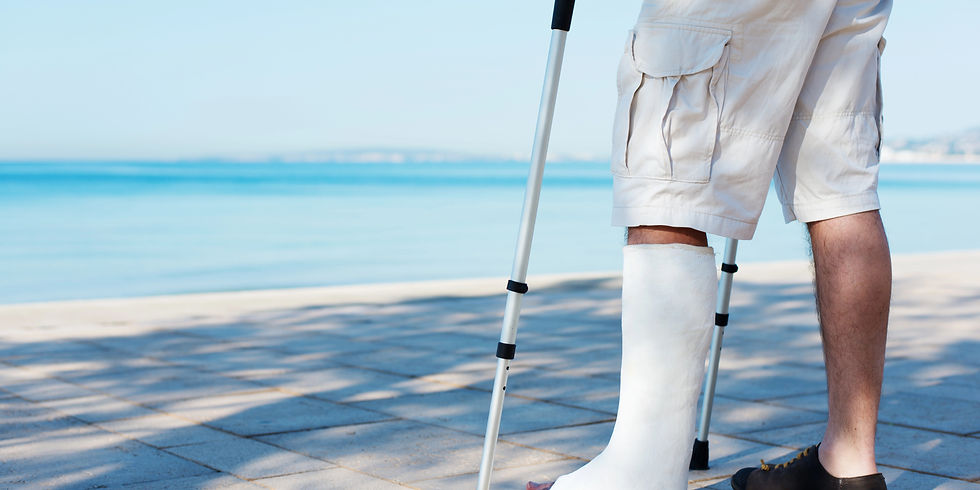 Man in shorts with a leg cast and crutches stands on a sunny waterfront promenade. The background features a calm blue sea.