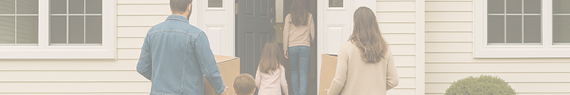 A family carrying boxes enters a white house, suggesting moving in. The mood is optimistic. Green shrubs are visible by the entrance.
