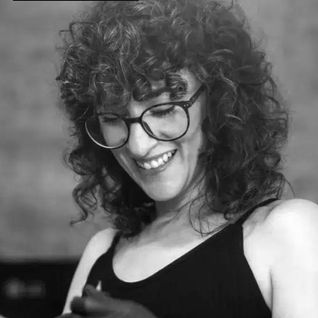 Mujer sonriendo con gafas y pelo rizado, mostrando tatuaje y guantes negros