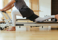 clinical pilates reformer single thigh stretch - holding onto bar, patient slides carriage back to stretch right hip flexors and quadricep muscles