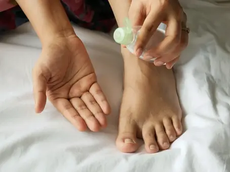 Hands applying Pitta pacifying oil to foot on white bedsheet. The person pours liquid from a small bottle into the palm, creating a calm, self-care mood.